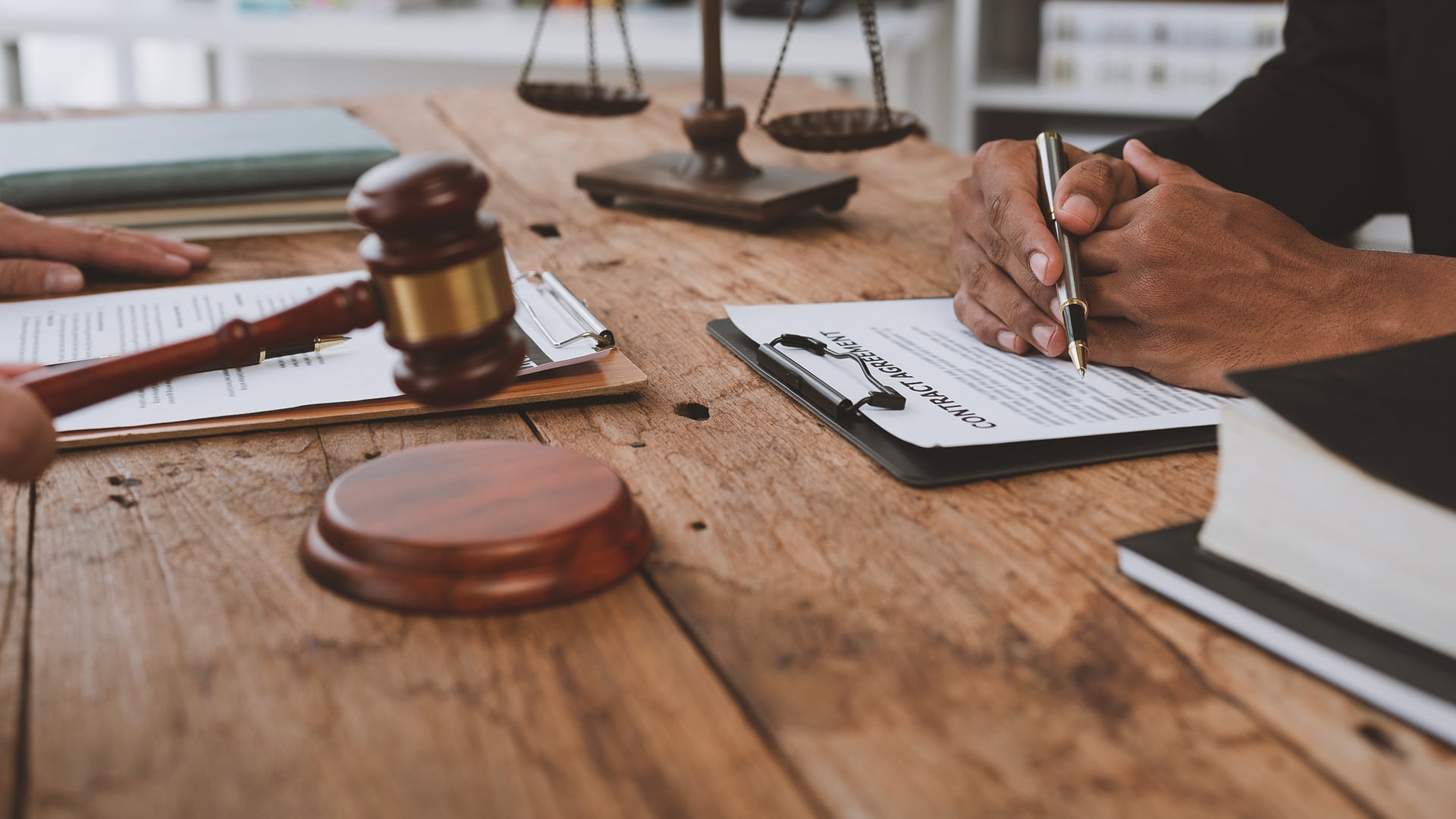 A close-up image of a gavel and a person's hands resting on a table with documents, a clipboard and some scales implying a decision is being made.