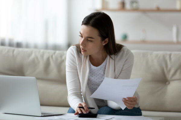 Caucasian woman with dark hair holds a letter in her hand whilst looking at her laptop, with a concerned look on her face