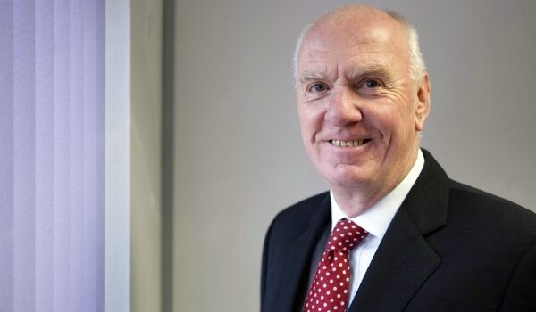 A smiling image Gerry Fitzjohn wearing a dark suit, white shirt and maroon tie.